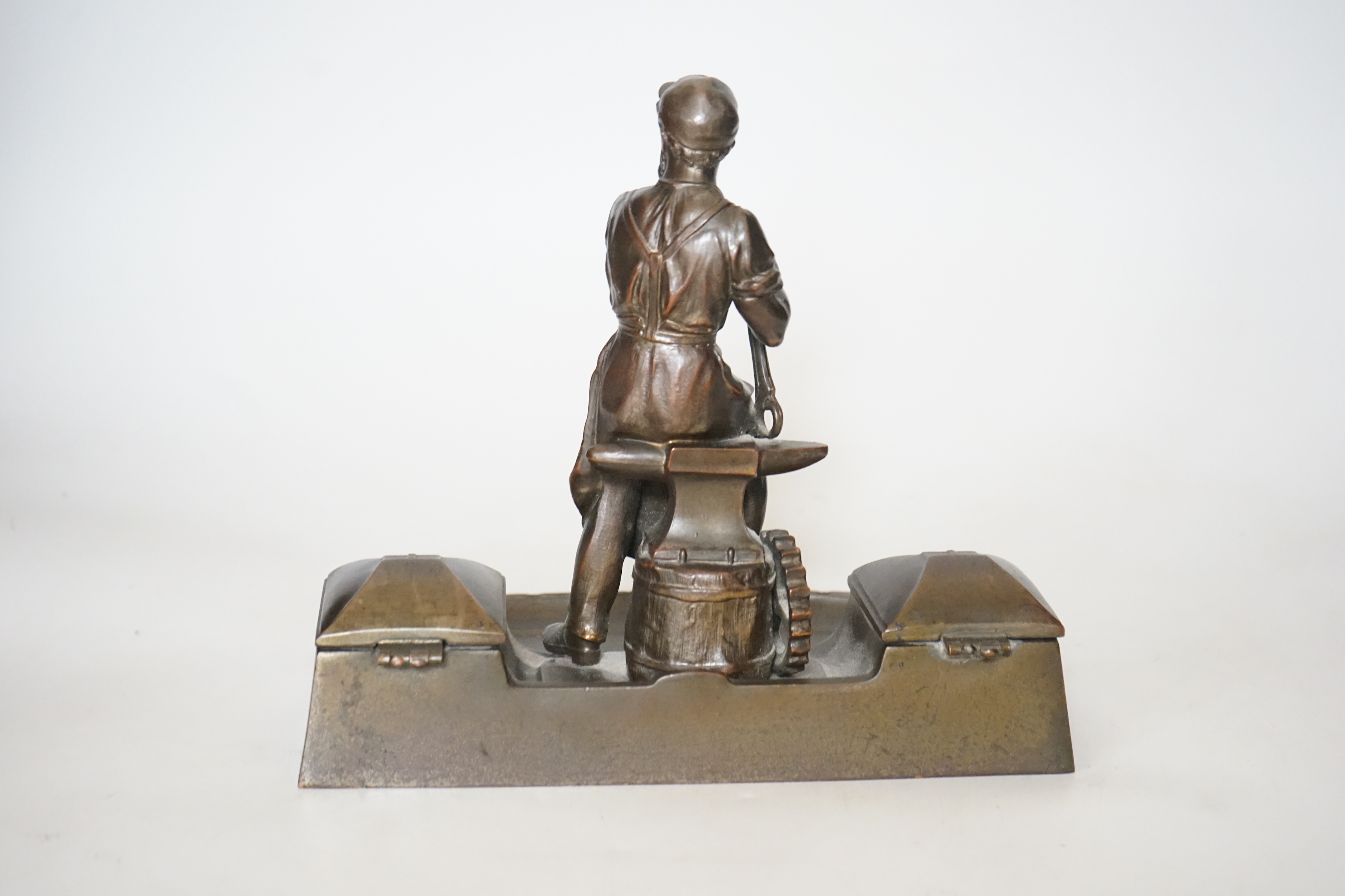 A WMF style spelter ‘Industry’ inkstand, figure with tools and an anvil, hinged ink wells either side, one glass ink bottle present, ‘1265’ cast into base, 20cm high